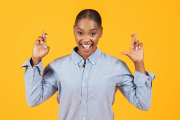 Joyful black woman crossing fingers making wish against yellow background