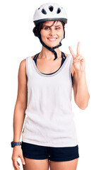 Beautiful young woman with short hair wearing bike helmet smiling with happy face winking at the camera doing victory sign. number two.