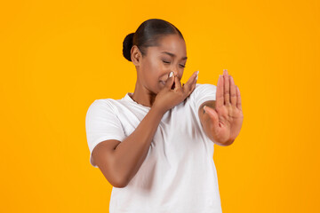 Disgusted young black lady closing nose showing STOP gesture, studio