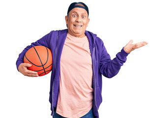 Senior handsome grey-haired man holding basketball ball celebrating victory with happy smile and winner expression with raised hands
