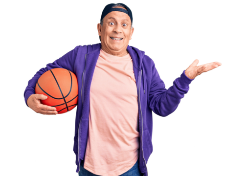 Senior handsome grey-haired man holding basketball ball celebrating victory with happy smile and winner expression with raised hands