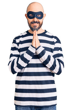 Young handsome man wearing burglar mask praying with hands together asking for forgiveness smiling confident.