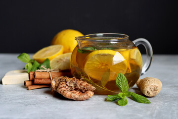 Fototapeta na wymiar Glass teapot with ginger tea, lemon, turmeric root and mint on grey background with space for text. Cold and flu season concept. 