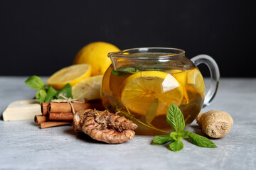 Glass teapot with ginger tea, lemon, turmeric root and mint on grey background with space for text. Cold and flu season concept. 
