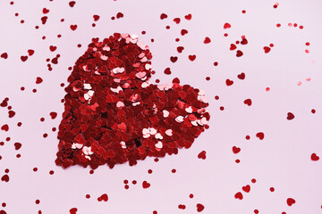 Heart made of shiny red small decorative hearts on a pink background strewn with sparkles. Side view, space for text. Valentine's Day card.