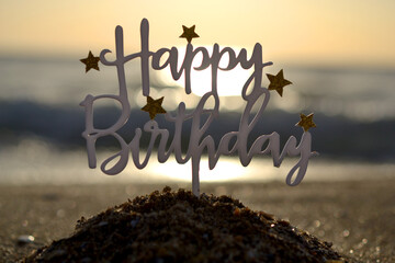 Stick in shape of letters Happy Birthday with stars at dawn and sunset on background of sea waves...