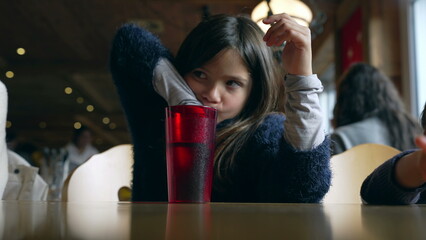 Small girl seated at restaurant waiting for food to arrive, drinks tap water from red plastic cup...