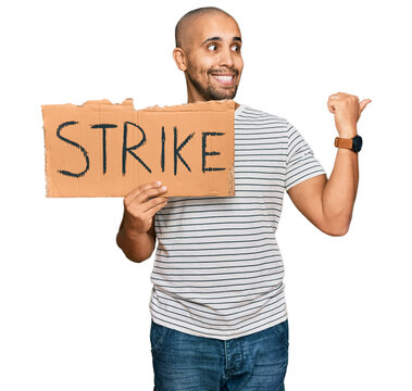 Hispanic adult man holding strike banner cardboard pointing thumb up to the side smiling happy with open mouth