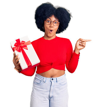 Young african american girl holding gift surprised pointing with finger to the side, open mouth amazed expression.