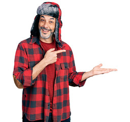 Middle age hispanic man with grey hair wearing fluffy earmuff hat amazed and smiling to the camera while presenting with hand and pointing with finger.