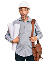 Middle age grey-haired man wearing safety helmet holding blueprints scared and amazed with open...