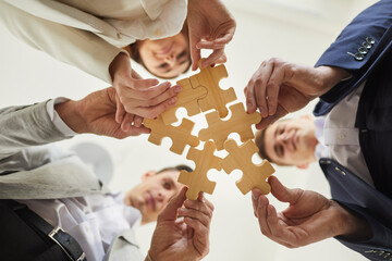 Business team trying to find solution to problem together. Group of young people holding parts of jigsaw puzzle. Crop shot close up bottom view from below hands holding wooden pieces. Teamwork concept - obrazy, fototapety, plakaty