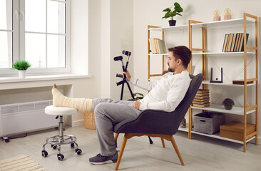 Man is sitting at home in chair near window with stand for broken bandaged leg and holding crutches in his hands. Recovery time after physical injury of foot bone. Orthopedic rehabilitation concept. - obrazy, fototapety, plakaty