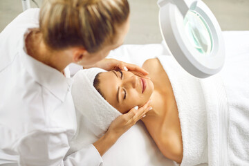 Young professional woman cosmetologist examining skin condition to a smiling girl client in beauty clinic. Beautician making facial massage or skin care procedure for treatment in spa salon.