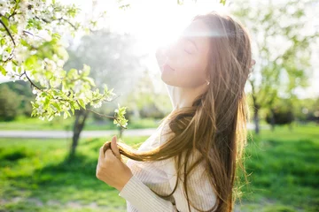 Tuinposter A close-up captures a lovely girl with flowing long hair posing amidst the blossoming trees of a spring park at sunset. It embodies the concepts of femininity, happiness, and love. © Konstantin Zibert