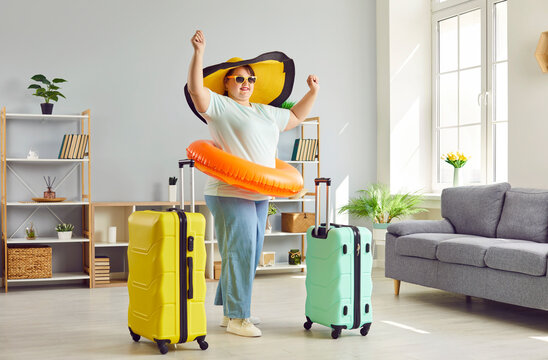 Funny, happy, fat woman, ready for summer vacation trip, wearing sun hat, swim ring and sunglasses, standing in living room with two suitcases, raising hands up and smiling. Holiday travel concept