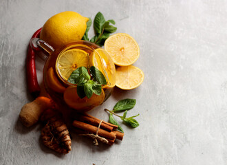 Glass teapot with ginger tea, lemon, turmeric root and mint on grey background with space for text. Cold and flu season concept. 