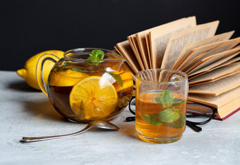 Glass teapot with ginger and lemon tea, open book and reading glasses on a table. Winter day still life. Healthy eating concept. 