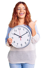 Young latin woman holding big clock pointing thumb up to the side smiling happy with open mouth