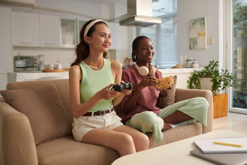 Smiling girl eating popcorn and watching her friend playing video game