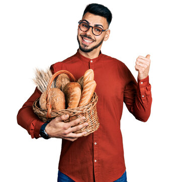 Young hispanic man with beard holding wicker basket with bread pointing thumb up to the side smiling happy with open mouth