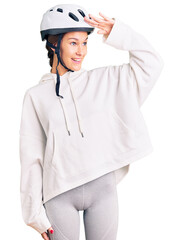 Beautiful brunette young woman wearing bike helmet and sporty clothes very happy and smiling looking far away with hand over head. searching concept.