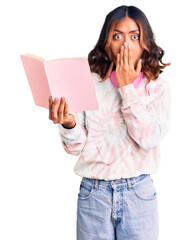 Young beautiful mixed race woman reading a book wearing headphones covering mouth with hand, shocked and afraid for mistake. surprised expression