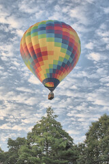 Tranquil early morning scene in New England countryside. Multicolored hot air balloon soaring over treetops and ascending into lovely blue sky with white puffy clouds.