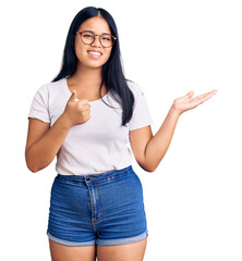 Young beautiful asian girl wearing casual clothes and glasses showing palm hand and doing ok gesture with thumbs up, smiling happy and cheerful