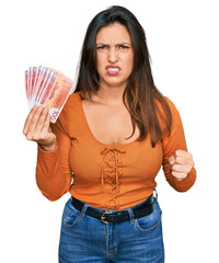 Beautiful hispanic woman holding norwegian krone banknotes annoyed and frustrated shouting with...