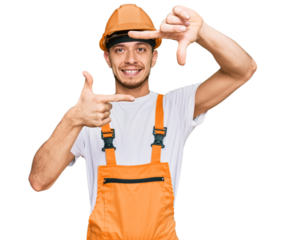 Hispanic young man wearing handyman uniform and safety hardhat smiling making frame with hands and fingers with happy face. creativity and photography concept.