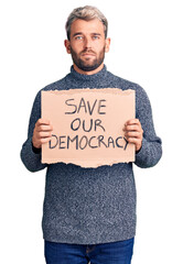 Young blond man holding save our democracy cardboard banner thinking attitude and sober expression...