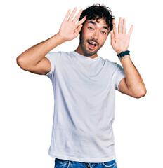 Fototapeta premium Hispanic young man with beard wearing casual white t shirt smiling cheerful playing peek a boo with hands showing face. surprised and exited