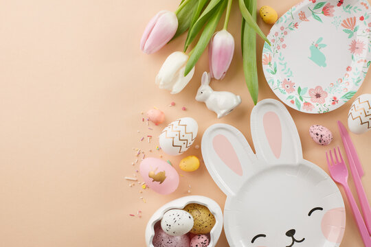 Children's Easter party concept. Top view photo of plate shaped like a rabbit, other plates, pink cutlery, painted eggs, fresh tulips, easter bunny on beige background with greeting space
