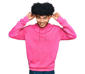 Fototapeta na wymiar Young african american man with afro hair wearing casual pink sweatshirt smiling pulling ears with fingers, funny gesture. audition problem