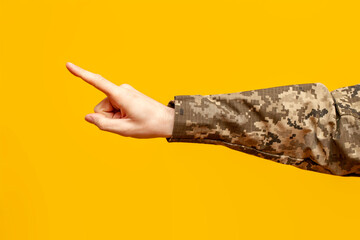 hand of a Ukrainian military man in camouflage uniform points with a finger and selects on a yellow isolated background, a male soldier points and presses, close-up - obrazy, fototapety, plakaty