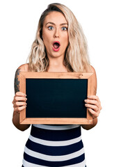 Young blonde woman holding blackboard afraid and shocked with surprise and amazed expression, fear and excited face.