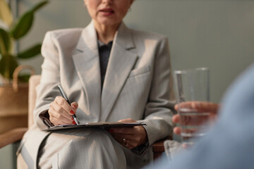 Close up of mature female therapist writing on clipboard while consulting unrecognizable client in office, copy space