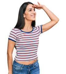 Young hispanic girl wearing casual striped t shirt very happy and smiling looking far away with hand over head. searching concept.