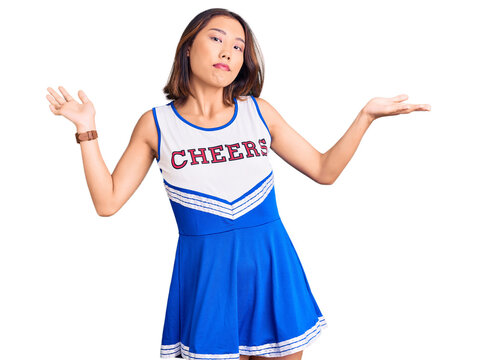 Young beautiful chinese girl wearing cheerleader uniform clueless and confused expression with arms and hands raised. doubt concept.