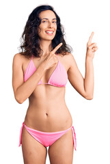 Young beautiful hispanic woman wearing bikini smiling and looking at the camera pointing with two hands and fingers to the side.