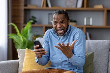 Close-up portrait of an angry African-American man sitting on the sofa at home, holding a mobile...