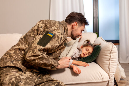young soldier of the Ukrainian army in a camouflage uniform returned home to his child, a military father covers and kisses his little daughter on the sofa at home, the concept of a military Ukrainian