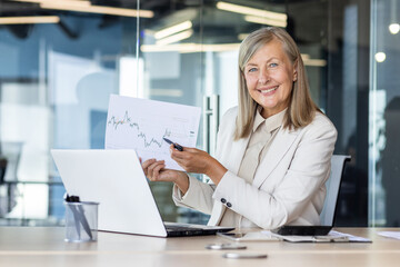 Portrait of a smiling senior successful business woman sitting in the office at the table, holding...