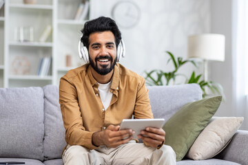 Portrait of a young Indian man wearing headphones sitting on the couch at home and holding a...