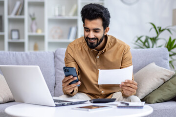 Fototapeta premium Smiling young Indian man sitting on sofa at home, working with documents, holding papers and using mobile phone.