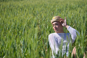 Naklejka na ściany i meble Non-binary person, young and South American, very makeup, wearing a hat, posing sitting in the middle of a field of green wheat. Concept queen, lgbtq+, pride, queer.