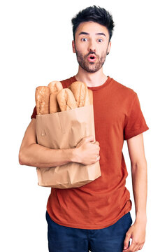 Young hispanic man holding paper bag with bread scared and amazed with open mouth for surprise, disbelief face