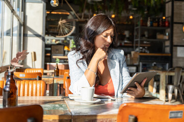 Woman looking at a digital tablet with a thoughtful expression while sitting in a coffee shop.