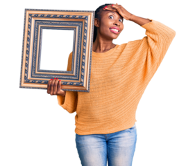 Young african american woman holding empty frame stressed and frustrated with hand on head, surprised and angry face